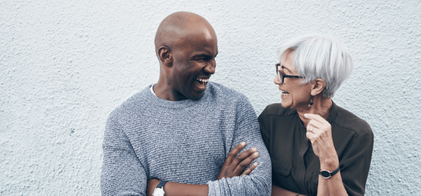 A black man and an older white woman laugh together while standing next to a wall.