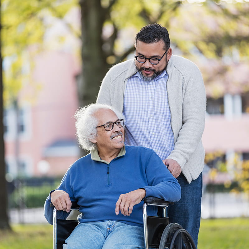 A man pushes an older man seated in wheelchair through a park. Both men are talking.