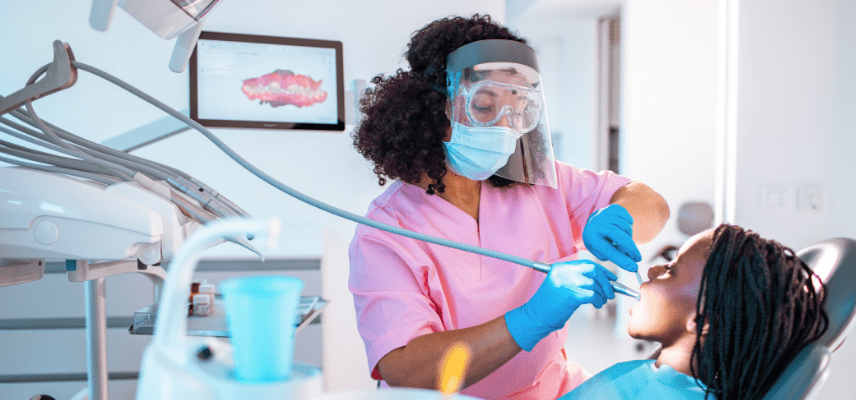 A black girl sits back in a dental chair as a black woman wearing dental clothing, gloves, and a face shield holds dental tools in the girl's mouth.