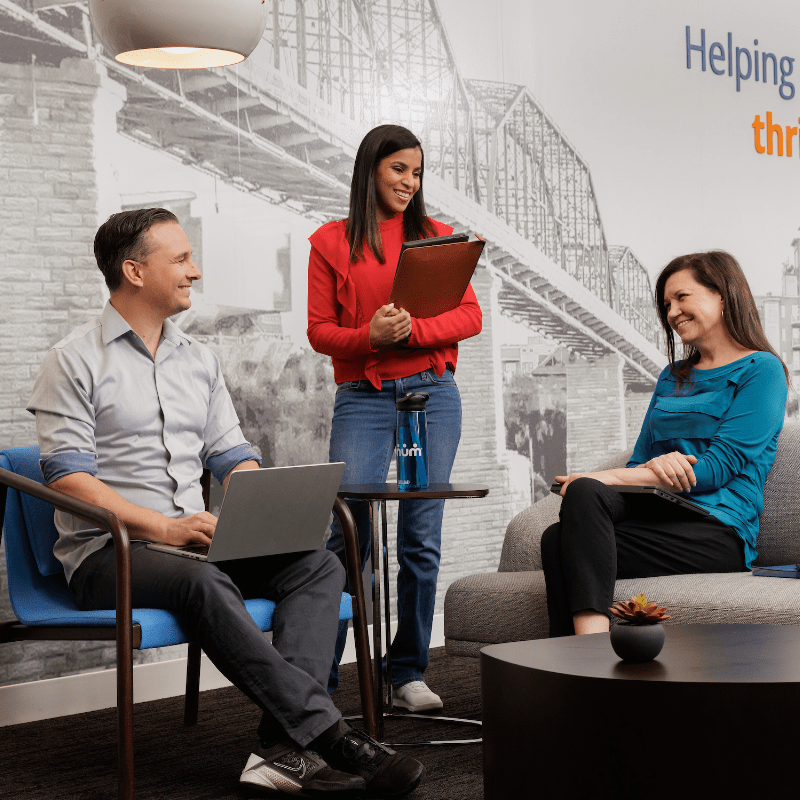A man and a woman discuss work with another woman seated in an office common area.