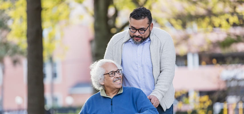 A man pushes a wheelchair used by an older man through a park. Both men are talking.
