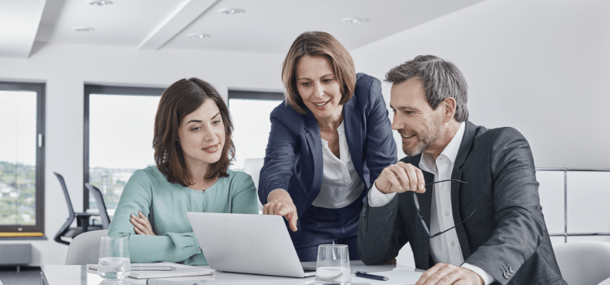 Three coworkers look at a laptop screen on an office table during a meeting. Two are seated and the third is standing between them, leaning forward, and pointing at the screen.