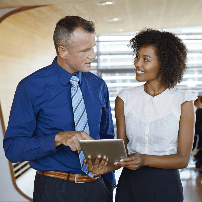 A white man points on a tablet device being held by a black woman as they're talking. They're standing in a conference room. Behind them are other individuals sitting at a conference table.