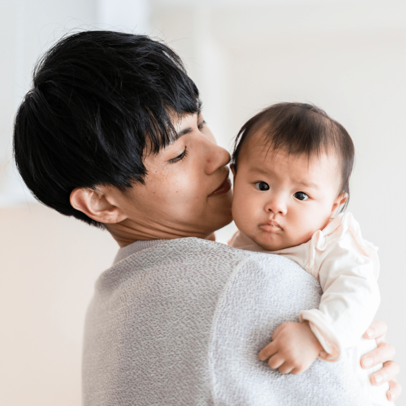 An Asian woman holds her infant child in her arms with her face next to the child's ear. The child's head is turned facing the camera.