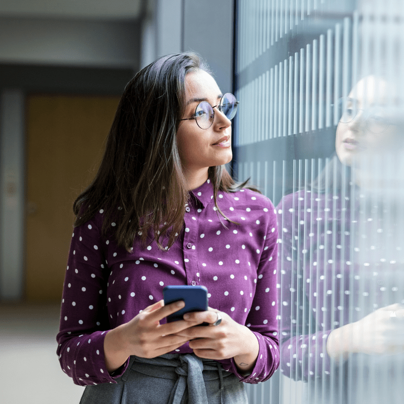 A woman looks out the window of hallway while holding a mobile device. The woman's reflection in the window is visible.
