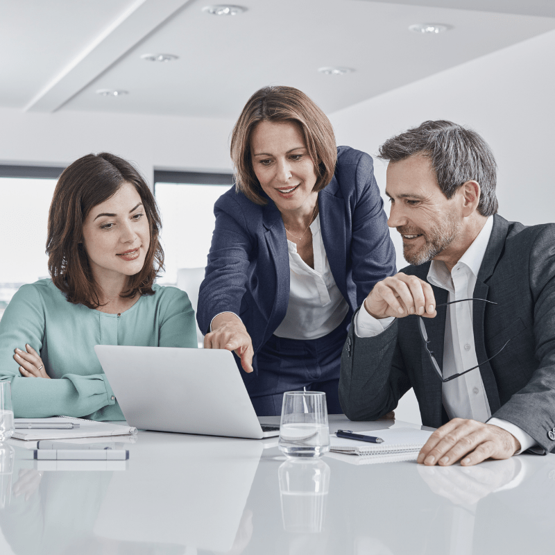 Three coworkers look at a laptop screen on an office table during a meeting. Two are seated and the third is standing between them, leaning forward, and pointing at the screen.